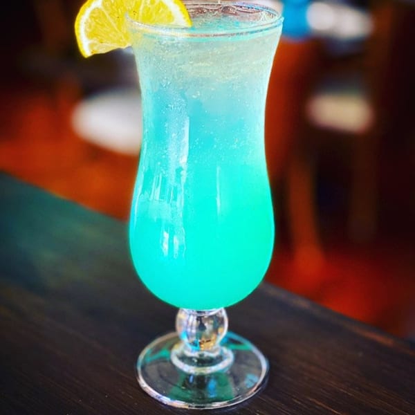A vibrant blue cocktail in a hurricane glass, garnished with a lemon slice, on a dark wooden table.