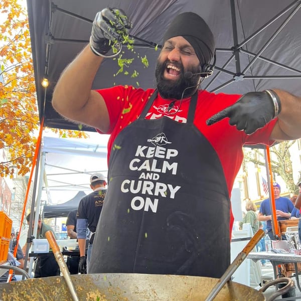 Aman, the chef, joyfully sprinkles fresh herbs into a large Indian curry pot at an outdoor event.