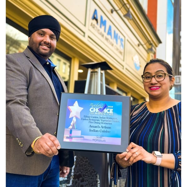 Two people proudly hold a "Reader's Choice 2024" award for Aman's Artisan Indian Cuisine outside the restaurant.